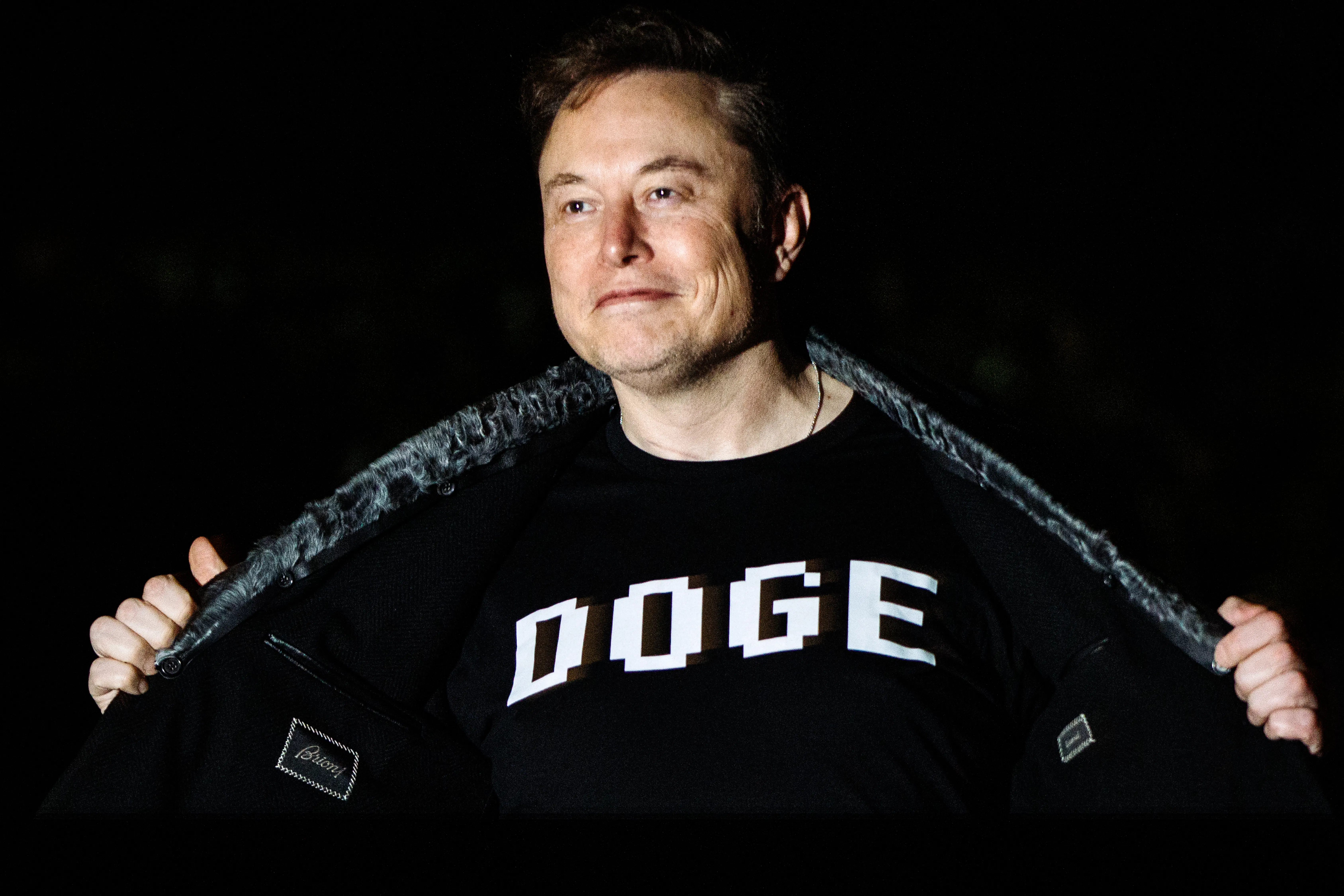 Elon Musk holds open his jacket to reveal a shirt that says "DOGE"