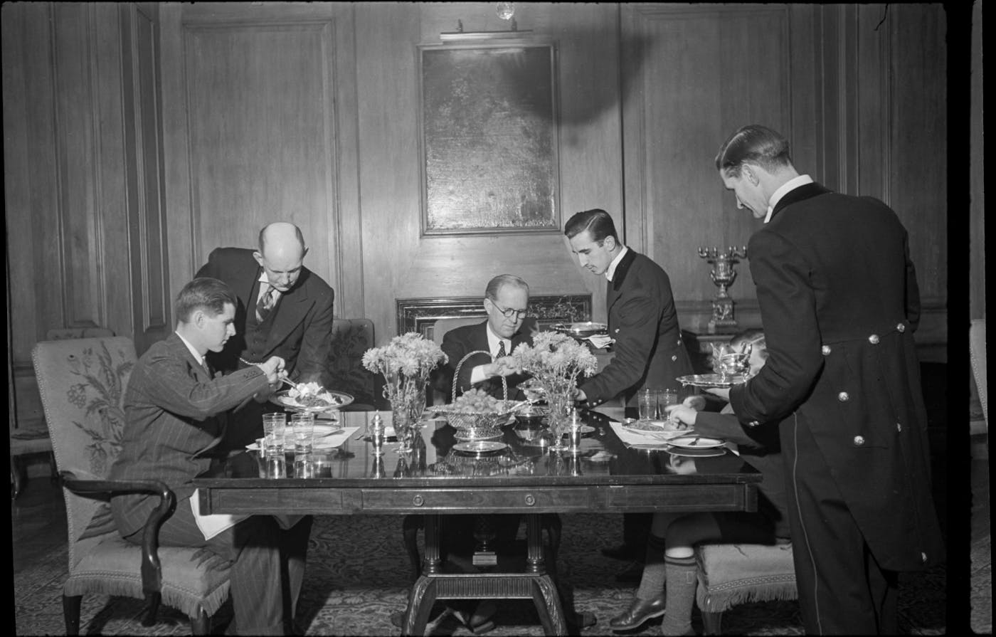 FDR-backing oligarch Joseph Kennedy is served dinner at a beautifully set table with three uniformed valets and his two children, including a young Teddy who is partially obscured behind the waiter on the right, but showing his knee high socks under the table. 