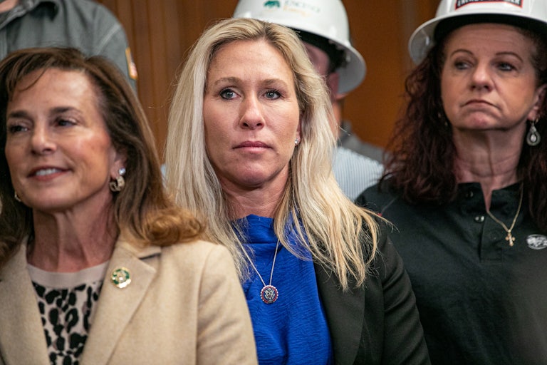 Marjorie Taylor Greene stands with a grim face. Others stand around her, a couple in hard hats.