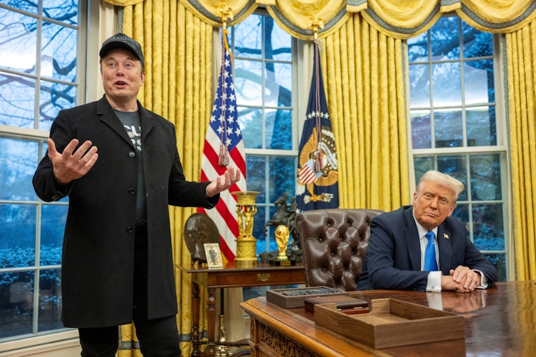 Elon Musk gestures and speaks in the Oval Office while Donald Trump sits at his desk