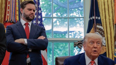 JD Vance stands with his arms crossed and looks down at Donald Trump, who sits at his desk in the Oval Office