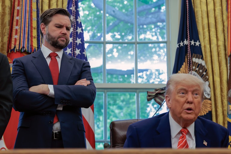 JD Vance stands with his arms crossed and looks down at Donald Trump, who sits at his desk in the Oval Office