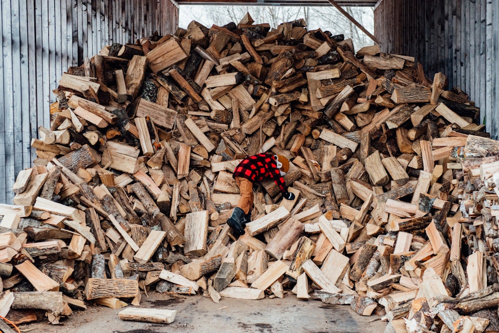 A child in boots and a plaid jacket climbs on an enormous pile of split wood in a partially open shelter.