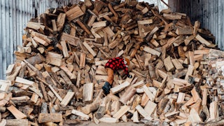 A child in boots and a plaid jacket climbs on an enormous pile of split wood in a partially open shelter.