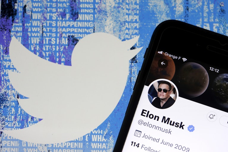 Elon Musk's Twitter account on a phone, with the Twitter logo (a blue bird) and the words "WHAT IS HAPPENING" on a screen behind it