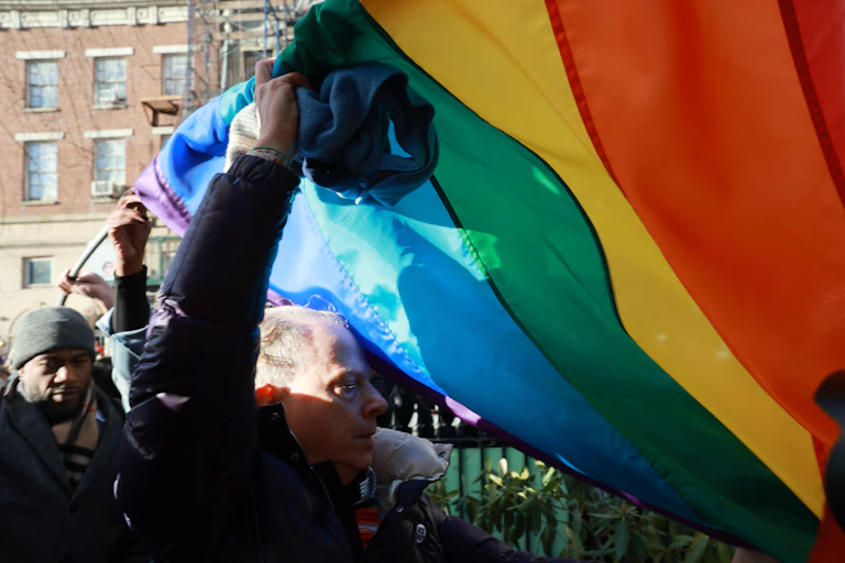Several people hold up a large Pride flag as others walk underneath