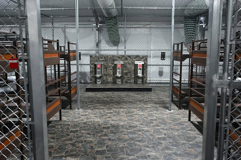 Three toilets side by side stand in the middle of a caged area with six bunk beds.