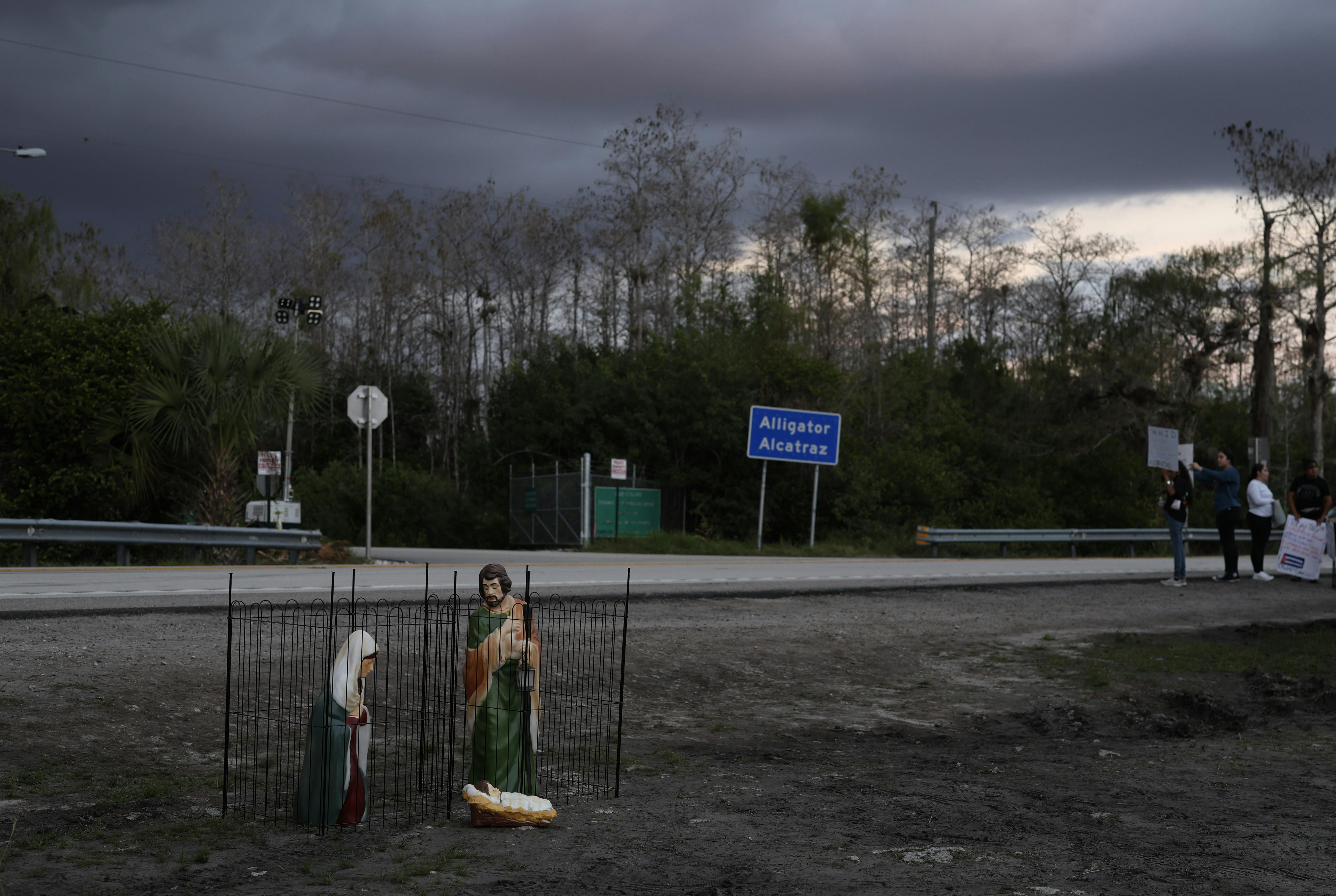  Nativity scene depicting the birth of Jesus Christ, featuring Mary and Joseph in cages as they are held in custody, sits near the entrance to "Alligator Alcatraz" as others protest in the background.