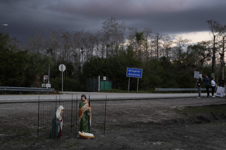 Nativity scene depicting the birth of Jesus Christ, featuring Mary and Joseph in cages as they are held in custody, sits near the entrance to "Alligator Alcatraz" as others protest in the background.