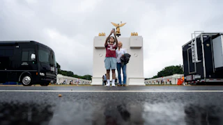 Two people take a selfie with a smartphone behind a puddle and in front of a white arch with gold statues on top.