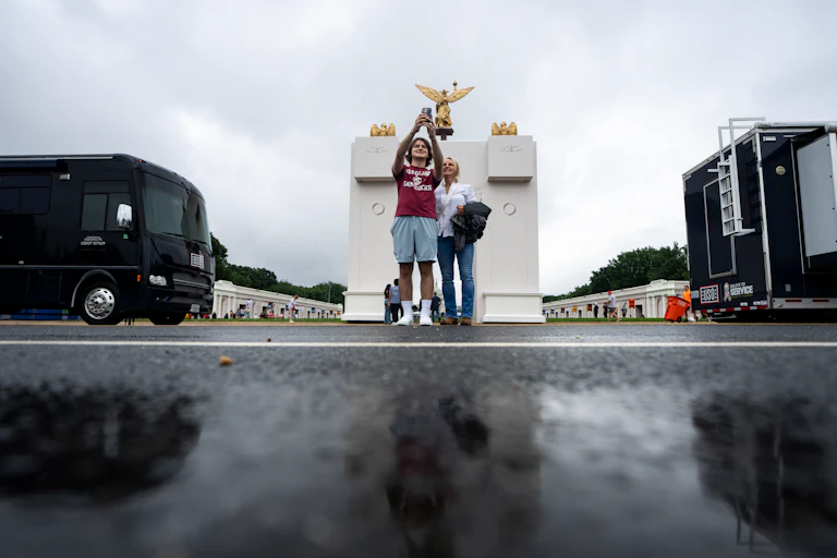 Two people take a selfie with a smartphone behind a puddle and in front of a white arch with gold statues on top.