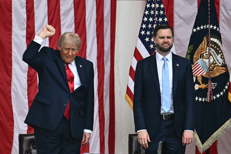 Donald Trump raises his fist and JD Vance stands with his arms hanging limply. A large U.S. flag is behind them.