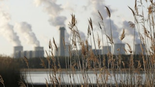 Grass is seen in the foreground with a lake beyond and long plumed from cooling towers on the other side of the lake.