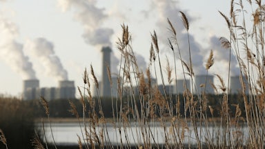 Grass is seen in the foreground with a lake beyond and long plumed from cooling towers on the other side of the lake.
