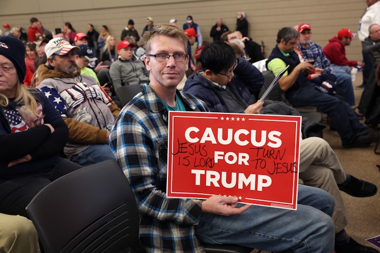 A man holds a sign that reads "Caucus for Trump." In handwritten words, it also reads "Jesus is Lord" and "Turn to Jesus."