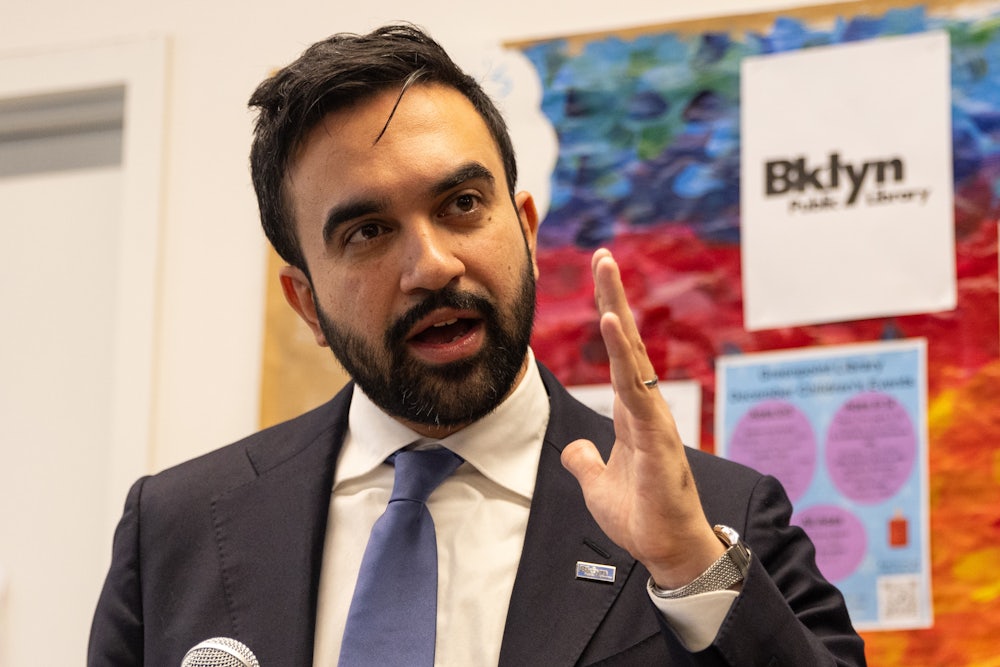 Mamdani holds up a hand while talking, in front of a colorful bulletin board.
