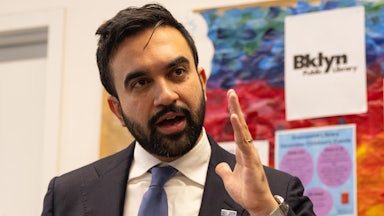 Mamdani holds up a hand while talking, in front of a colorful bulletin board.