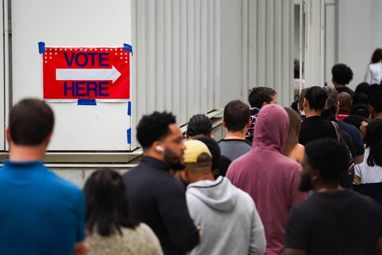 A long line of people. A sign next to them reads "Vote Here" with an arrow sign.