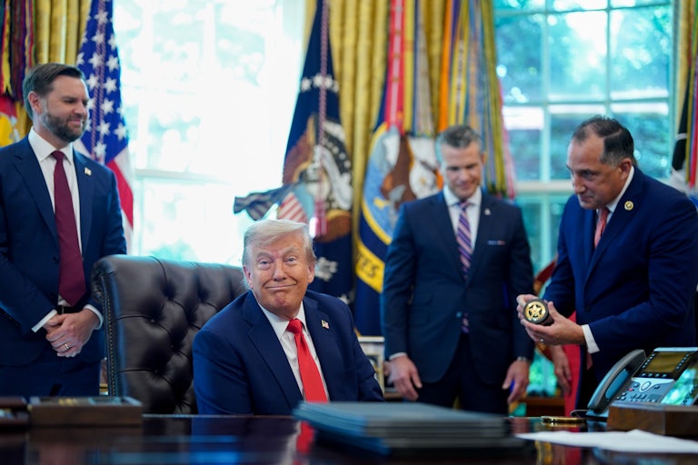 Donald Trump, seated at his desk in the Oval Office, smiles at the camera as Gadyaces Serralta, director of the US Marshals Service, bends down slightly holding a police badge in his hands. Pete Hegseth and JD Vance look on smiling.