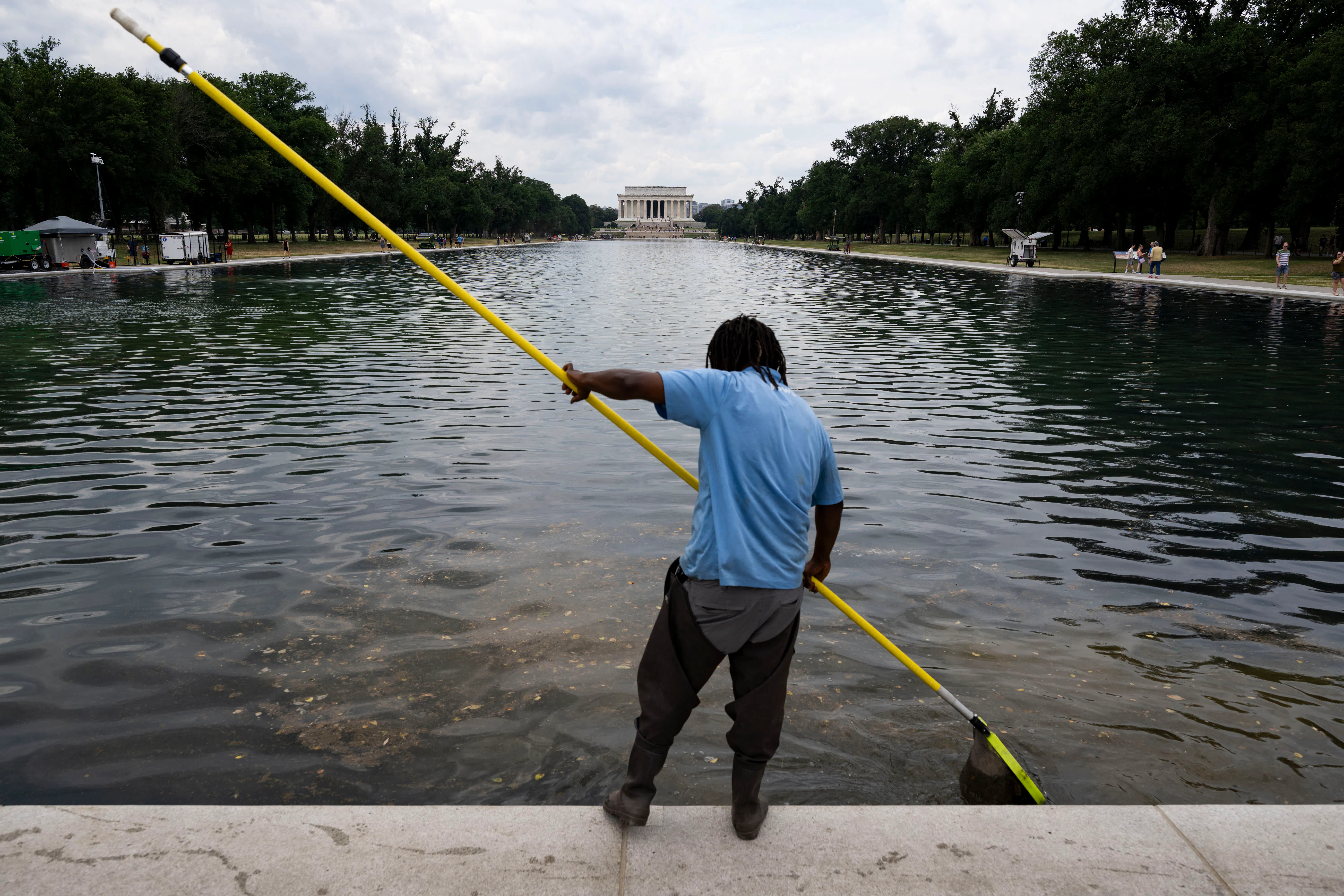 A man in a blue shirt and dark pants uses a long yellow pole to clean the Lincoln Memorial Reflecting Pool, with the memorial building visible in the background.