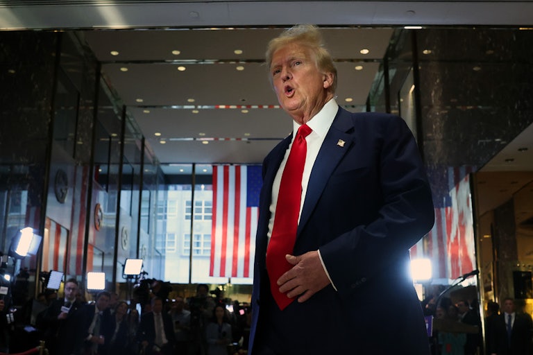 Donald Trump is yelling inside Trump tower. A crowd (dark, out of focus) is behind him as well as a U.S. flag draped from the ceiling in front of the window.
