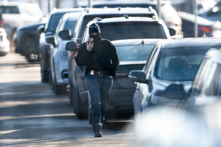 A masked ICE agent walks by some cars while he holds his phone up to his face.