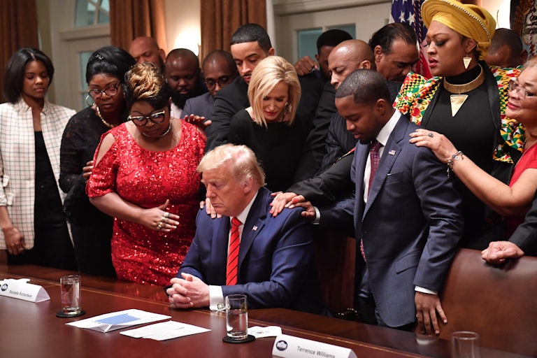 Donald Trump sits at his desk as more than a dozen African Americans stand around him, some placing their hands on his shoulders, heads bowed and eyes closed.