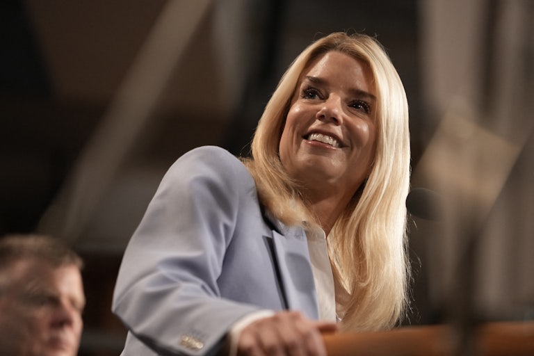 Attorney General Pam Bondi smiles and looks to the side while standing at a podium during a press conference