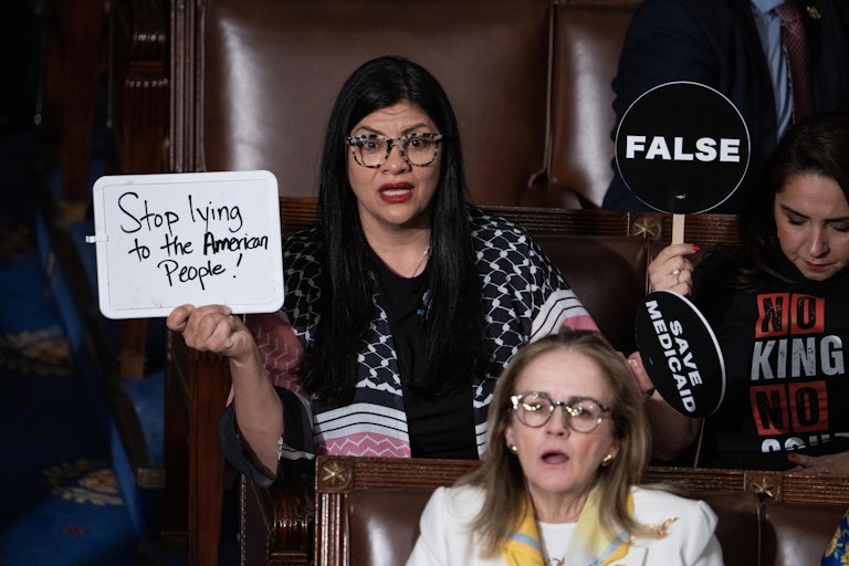 Representative Rashida Tlaib sits in the Capitol wearing a keffiyeh and holding up a whiteboard on which she wrote "Stop Lying to the American People!" She looks outraged. Others around her hold up signs that read "False" and "Save Medicaid."