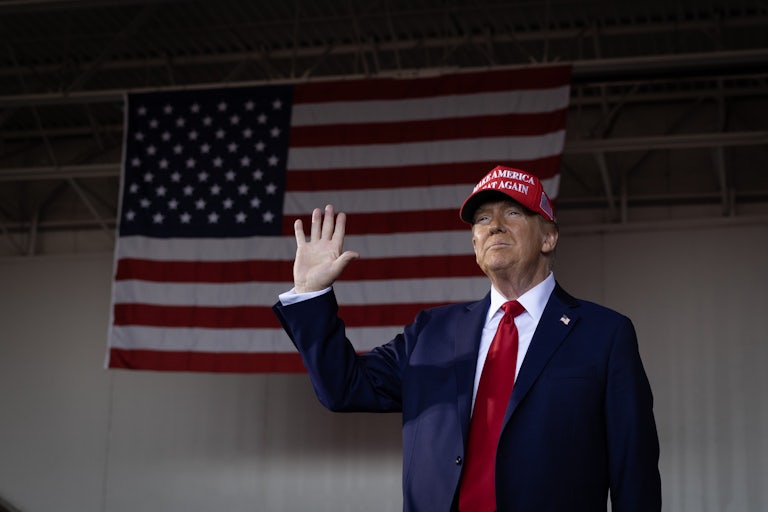 Donald Trump, wearing a MAGA hat, smiles and waves in front of a large U.S. flag hung on the wall.