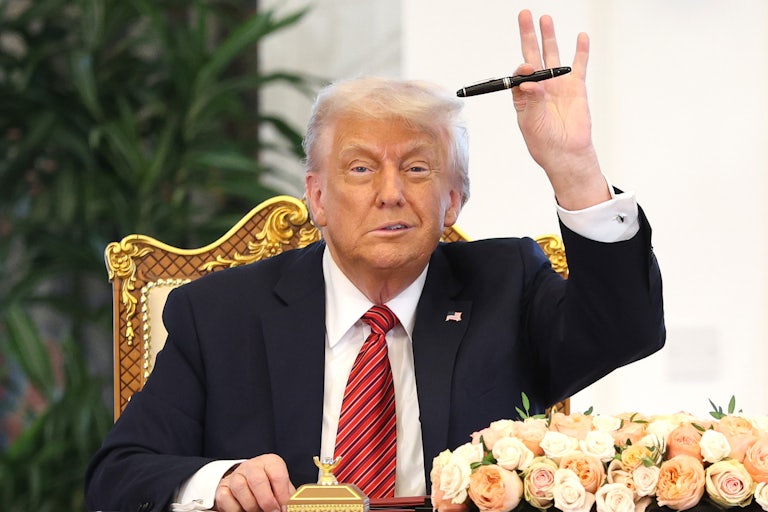 Donald Trump makes a weird face while waving with a pen on his hand. He sits on a gold chair.