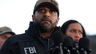 Kash Patel wears an FBI hat and FBI jacket while standing in front of a lectern with several microphones.