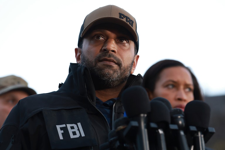 Kash Patel wears an FBI hat and FBI jacket while standing in front of a lectern with several microphones.