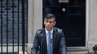 Rishi Sunak stands behind a podium as he gives a speech in the rain.