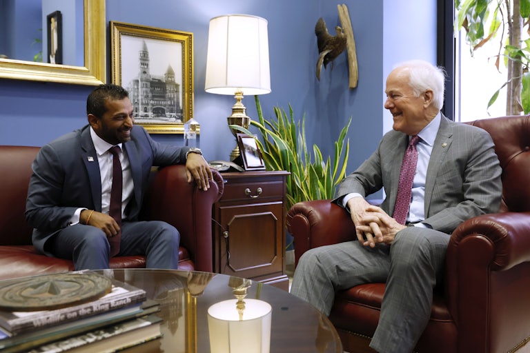 FBI Director Kash Patel and Senator John Cornyn are laughing while seated on a couch and armchair.