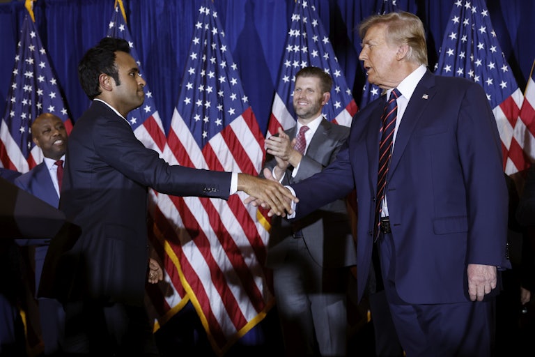 Vivek Ramswamy shakes hands with Donald Trump on a stage. Several U.S. flags, Eric Trump, and Tim Scott are in the background.