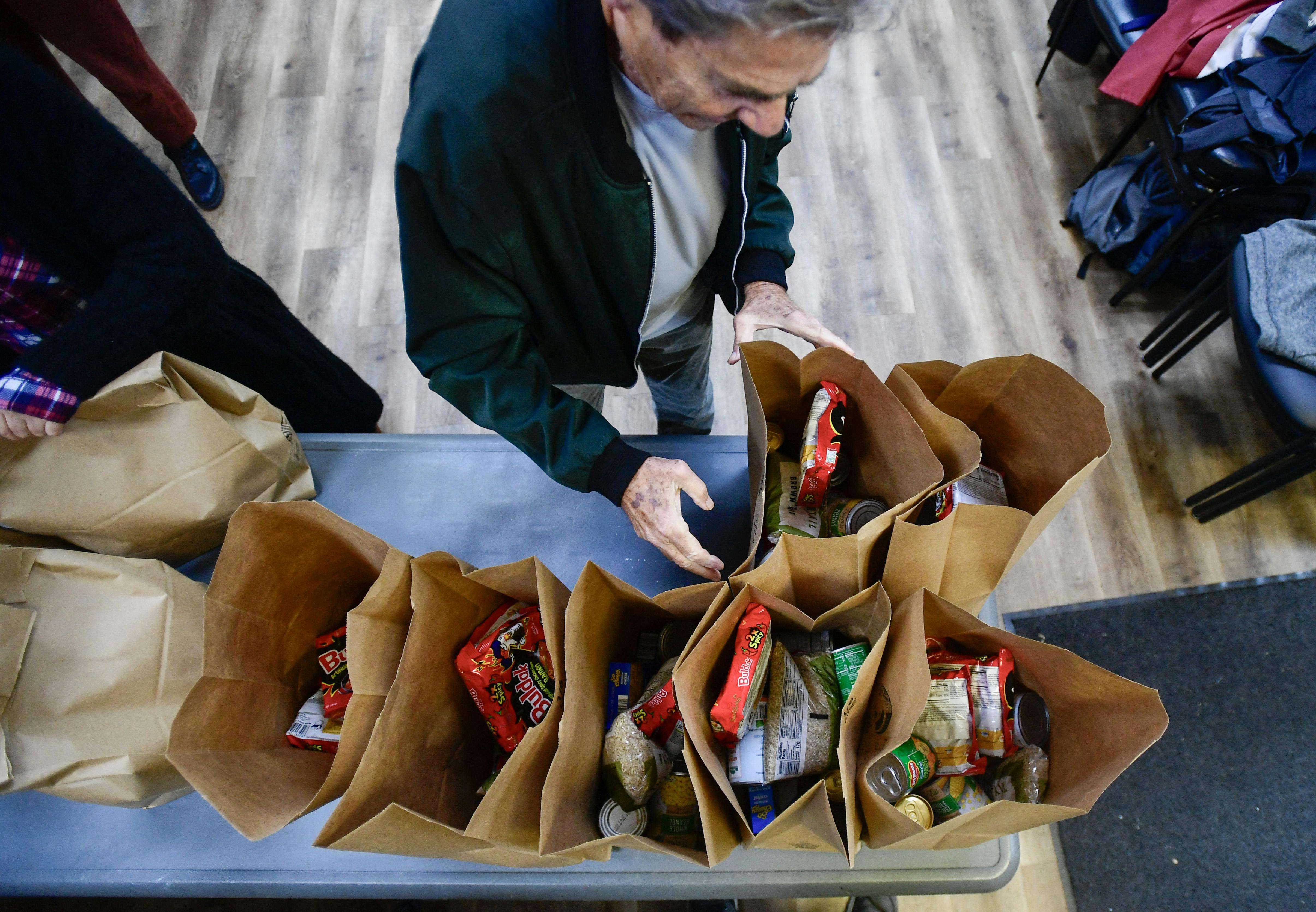 An overhead photo shows a person reaching towards paper bags filled with groceries, on a table.