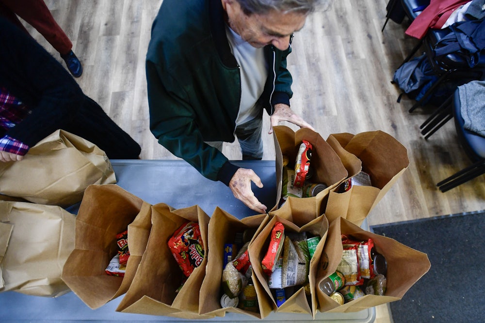 An overhead photo shows a person reaching towards paper bags filled with groceries, on a table.