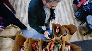 An overhead photo shows a person reaching towards paper bags filled with groceries, on a table.