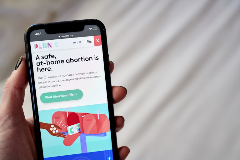 A hand with nail polish holds a smartphone with the Plan C website