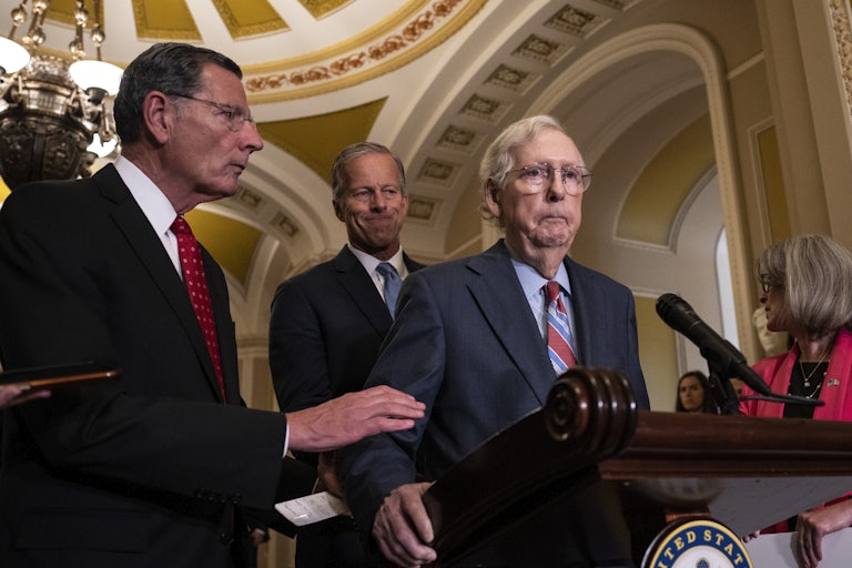 Mitch McConnell freezes at a lectern in the Capitol as other lawmakers try to stand near him looking concerned.