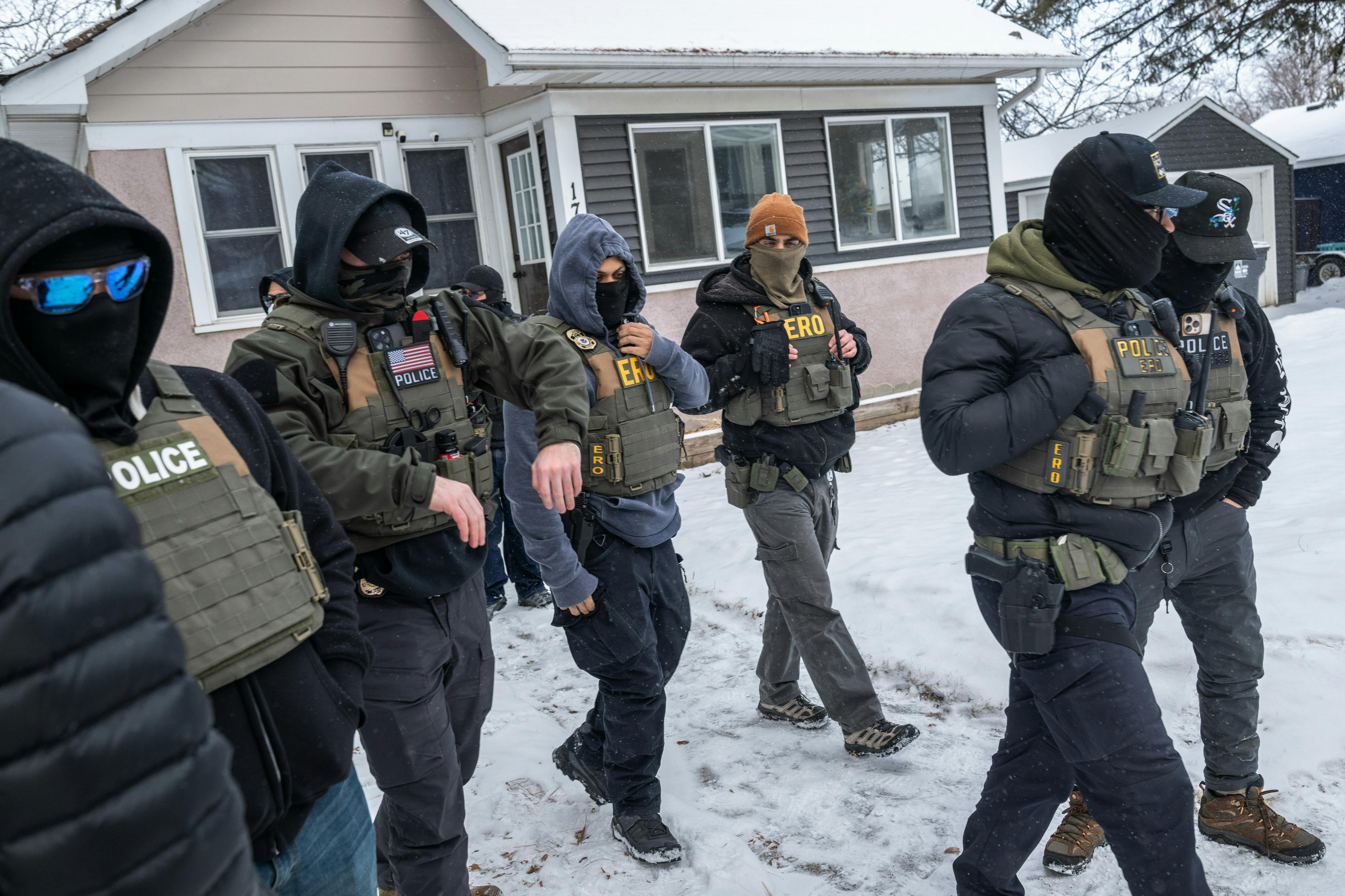 Masked federal agents walk outside a home in Minneapolis, the yard blanketed with snow.