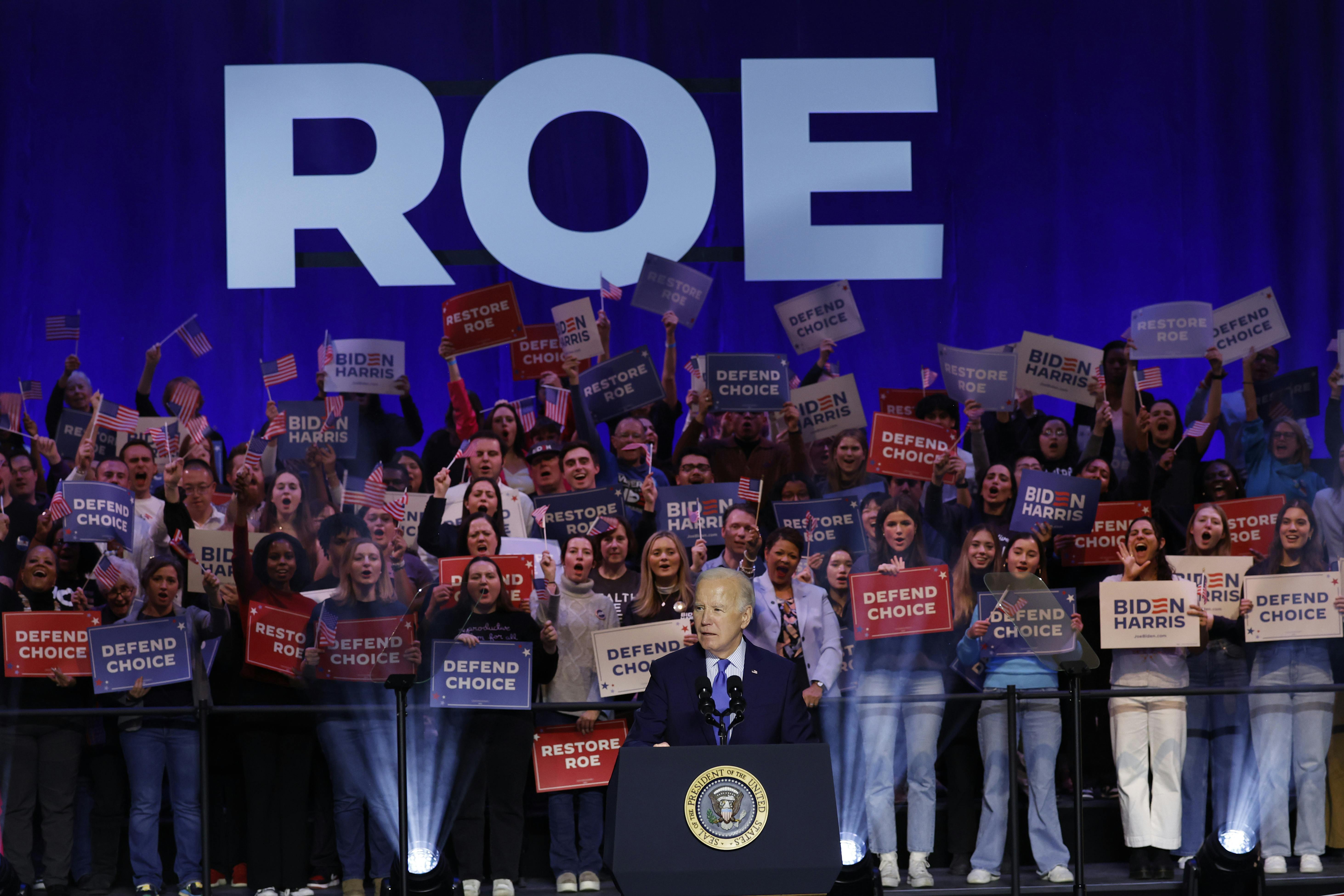 Biden stands at a lectern with the word "Roe" behind him and people holding signs saying "Defend Choice."