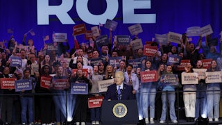Biden stands at a lectern with the word "Roe" behind him and people holding signs saying "Defend Choice."