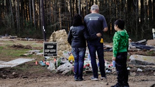 Three people stand at the site of a memorial, next to a sign reading "Stop Cop City."