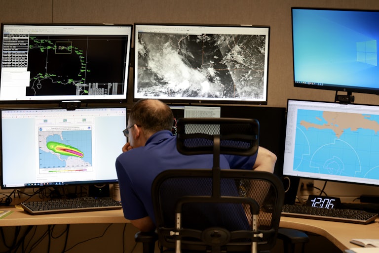 A man sits hunched over staring at one of six computer screens in front of him showing weather patterns.