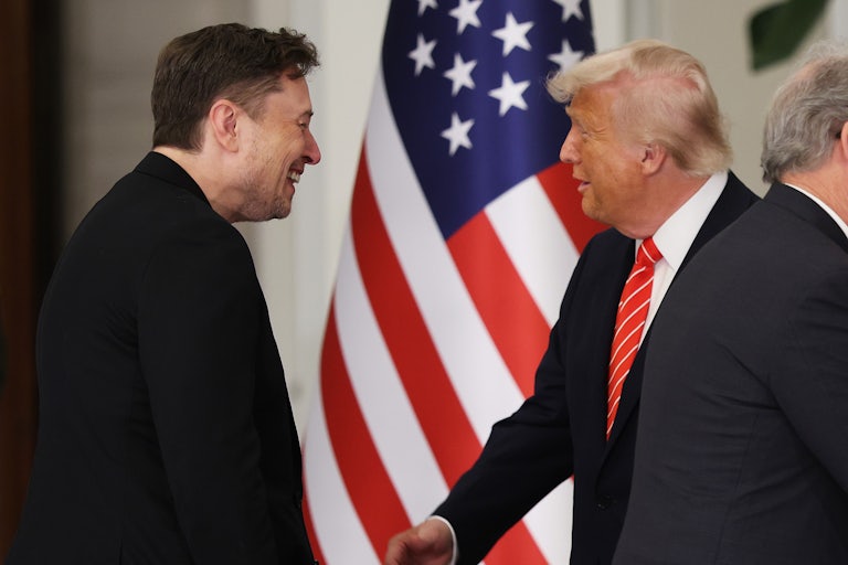 Donald Trump and Elon Musk laugh while standing in front of a U.S. flag.