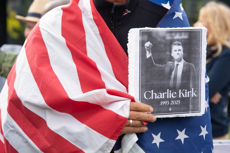 A person has an American flag draped around their shoulders and holds a photo of Charlie Kirk