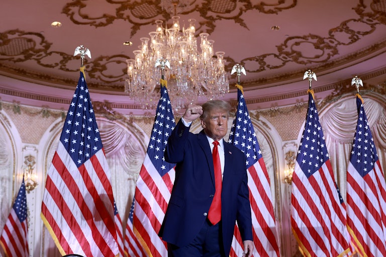 Donald Trump stands in front of a row of U.S. flags at Mar-a-Lago and raises a clenched fist as in victory.