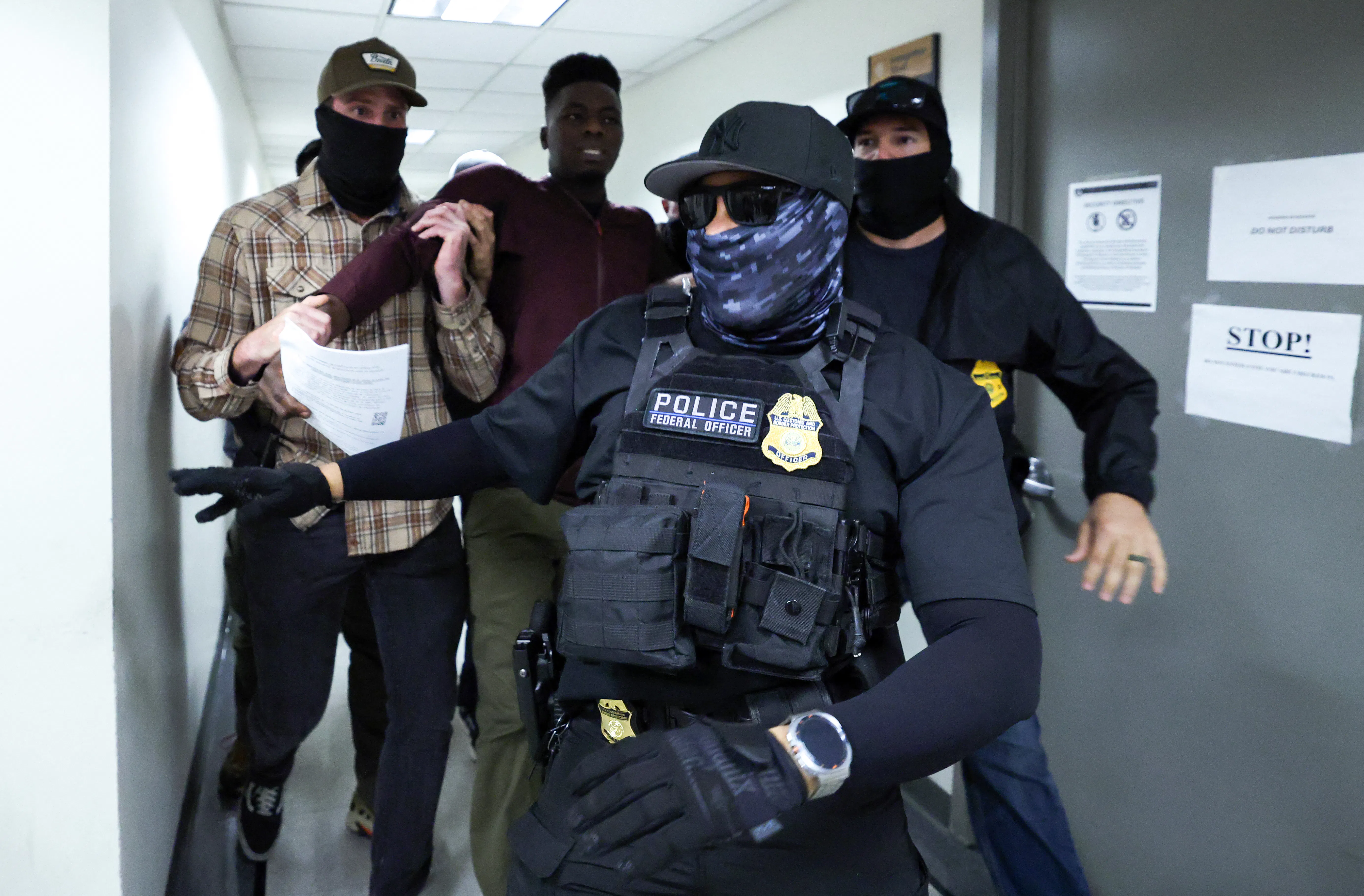 Two masked federal agents detain a man in the hallways of the Jacob K. Javitz Federal Building while another masked agent wearing a flak jacket and gloves walks ahead of them.